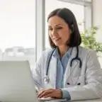 Smiling doctor using laptop in modern medical office to review clinic performance metrics and patient data.