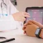 A doctor in a white coat is speaking with a patient seated across a desk, whose hands are clasped and wearing a smartwatch. A tablet on the desk displays health metrics like sleep tracking and heart rate in a dashboard format.
