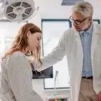 A middle-aged male doctor in a white coat gently consoles a young female patient in a hospital gown, seated on an exam table in a brightly lit medical room.