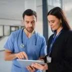 A clinician in scrubs and an administrator in professional attire look at a tablet together inside a bright medical facility.