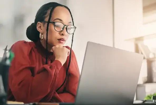 Woman wearing glasses sits at a desk using a laptop, focused on reviewing healthcare or medical billing information in a bright, modern workspace.
