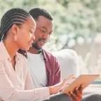 A couple sitting together on a couch, reviewing healthcare information on a tablet in a bright living room.
