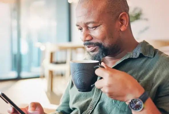 Middle-aged man sitting indoors, holding a coffee mug and looking at his smartphone in a bright, modern setting.